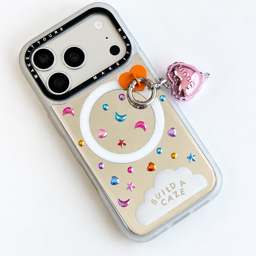 A mirror phone case with colorful rhinestones, crescent moon and star stickers, a white circular ring, and a pink heart-shaped charm engraved with 'BUILD A CAZE'.