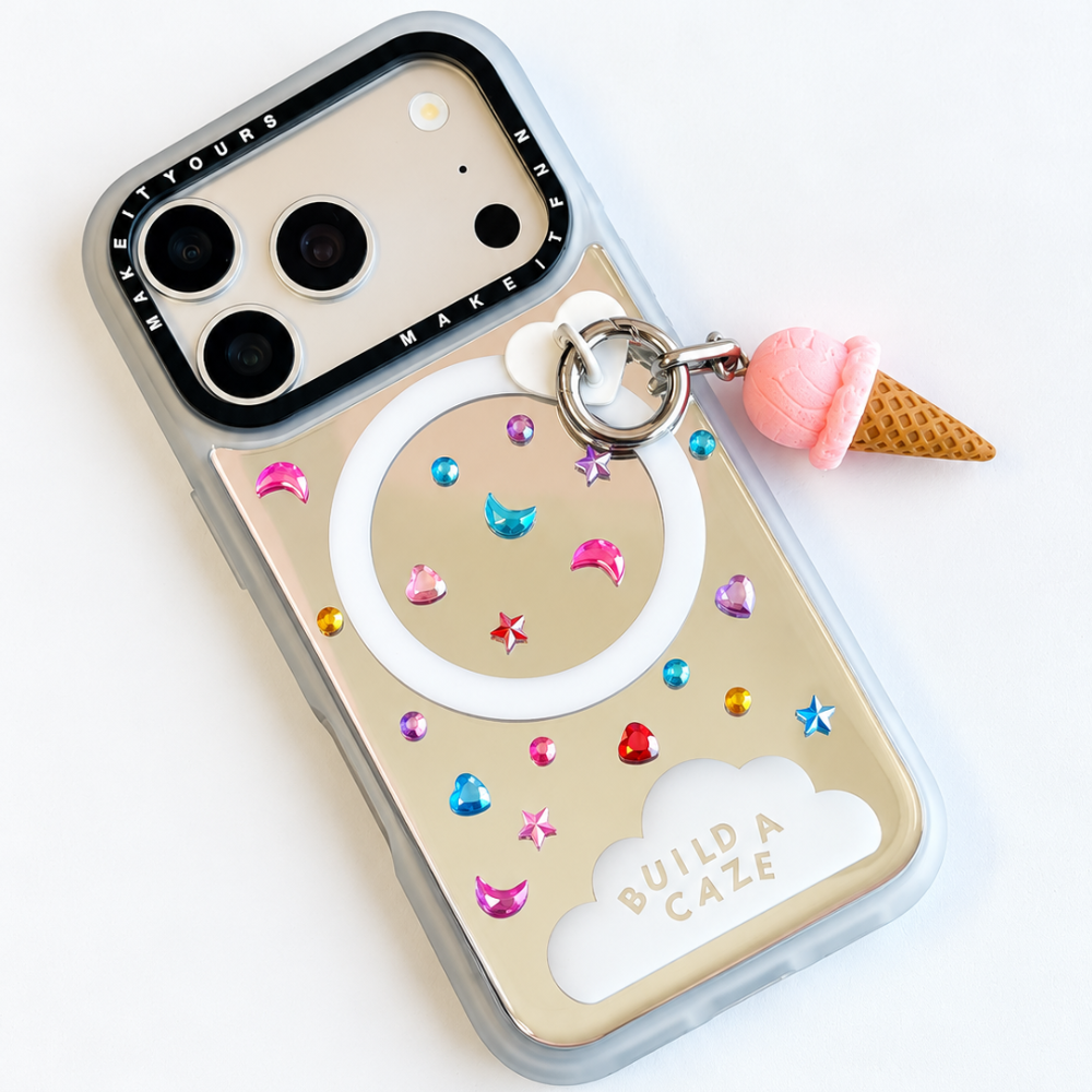 A mirror phone case with colorful rhinestones, crescent moon and star stickers, a white magsafe ring, an ice cream cone charm engraved with 'BUILD A CAZE'.