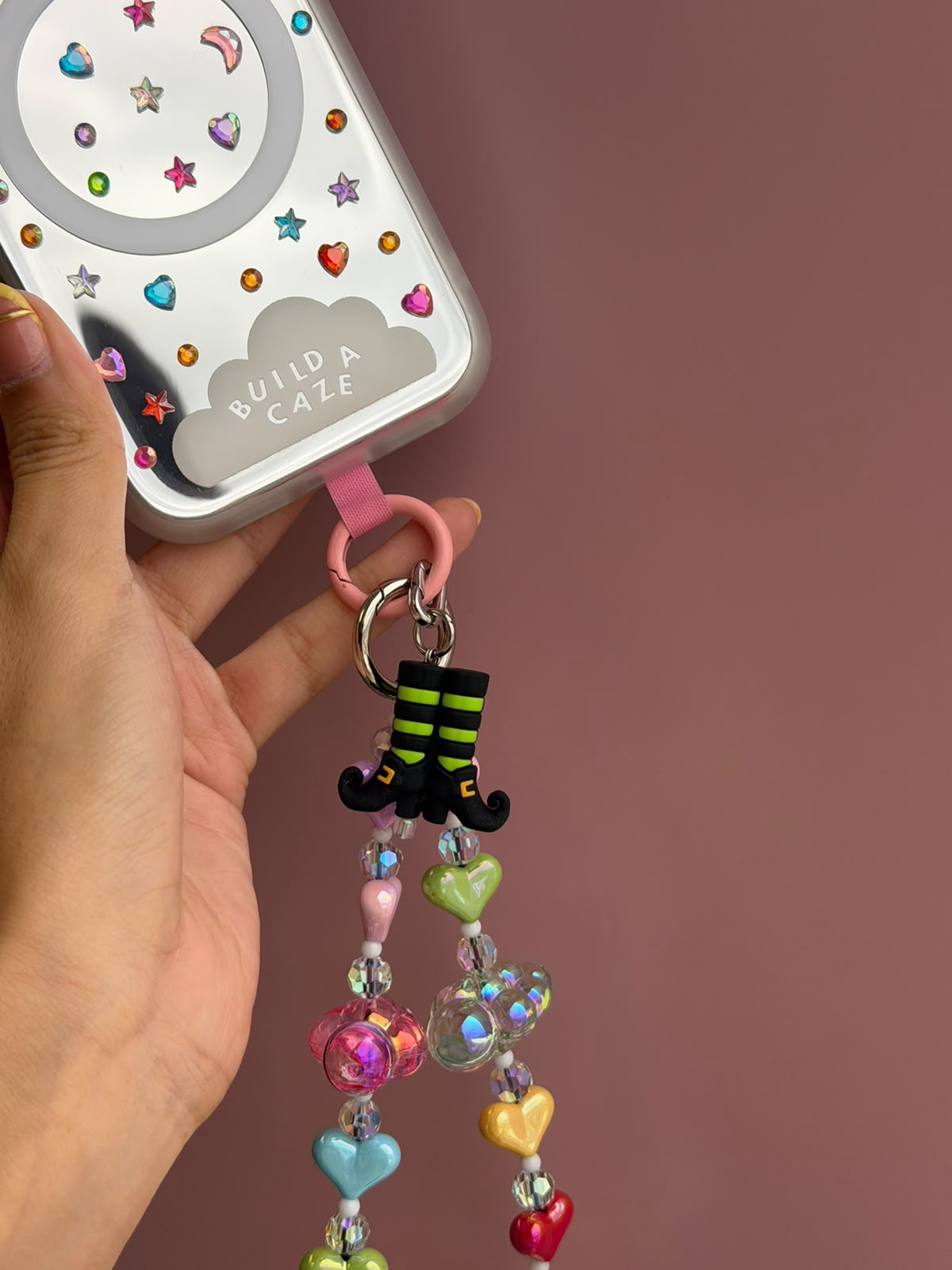 A hand holds a phone case decorated with colorful rhinestones and a pink 'BUILD A CAZE' cloud logo, attached to a pink keychain loop. The keychain features a black and lime green witch boot charm and dangling pastel heart beads on a dusty rose background.