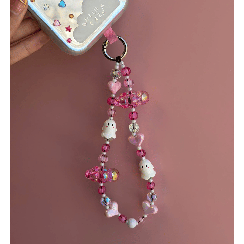 A pink and white beaded phone strap with cartoon ghost charms, heart beads, and iridescent crystal accents, shown attached to a phone case held by a hand against a dusty pink background.
