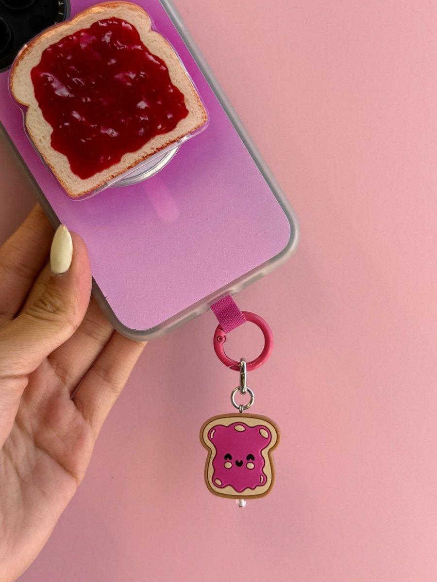 Hand holding a purple phone case with the Raspberry Pink Jammy Toastie Charmie clipped on, next to a slice of toast with jam